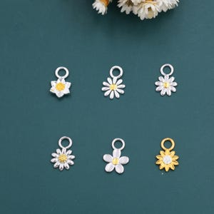 May include: Six daisy-shaped charms in silver and gold. Each charm has a loop for attaching to jewellery. The daisy petals are white, and the centres are yellow. The charms are displayed on a dark teal background, with some dried flowers in the top corner.
