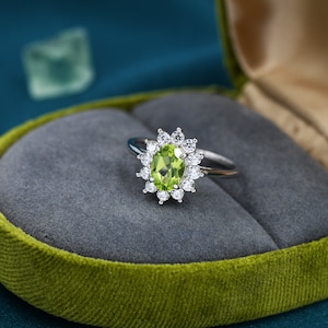 May include: A silver ring featuring a large green oval gemstone surrounded by smaller clear gemstones. The ring is displayed in a green velvet ring box.