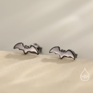 Puede incluir: Un par de pendientes de botón plateados con forma de murciélago. Cada pendiente presenta una silueta de murciélago con cuerpo blanco y alas gris oscuro. Los pendientes se muestran sobre una superficie beige claro.