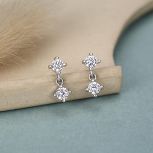 May include: A pair of silver-toned drop earrings, each featuring two round, clear, faceted stones set in a four-prong metal setting. The top stone is fixed, while the bottom stone hangs from a small circular link, creating a dangling effect.