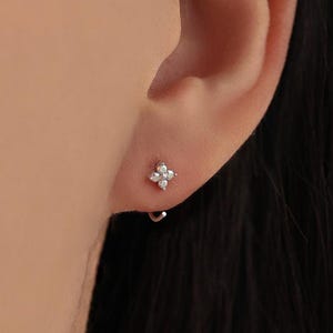 May include: A small, silver earring with a four-petal flower design, set with clear gemstones. The earring is shown on an ear, highlighting its delicate and minimalist style. The earring is a small hoop style.