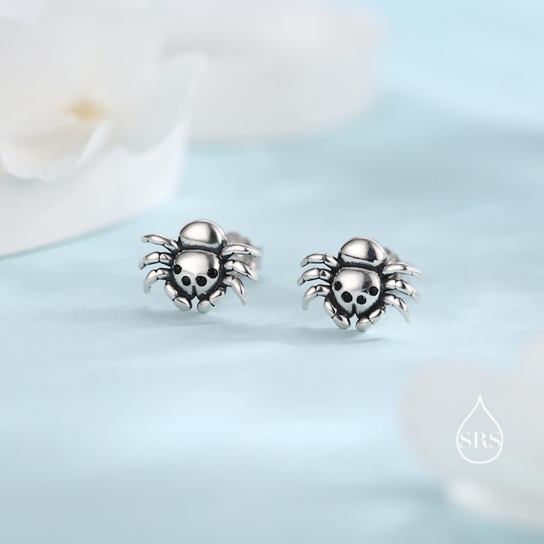 May include: A pair of silver spider stud earrings. The spiders are detailed and have black eyes.