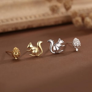 Può includere: Orecchini a lobo con design a scoiattolo e pigna, in tonalità oro e argento. Gli orecchini sono esposti su una superficie marrone.