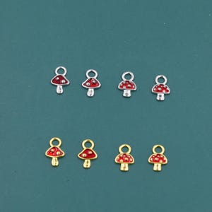 Puede incluir: Ocho dijes de champiñones con tapas rojas y lunares blancos. Los dijes tienen bases y bucles de metal plateado y dorado para sujetarlos a joyas u otras manualidades. Los champiñones están dispuestos en dos filas de cuatro.
