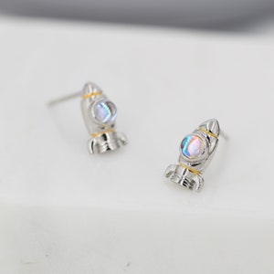 May include: A pair of silver rocket ship earrings with iridescent blue and white stones. The rocket ships have gold accents.