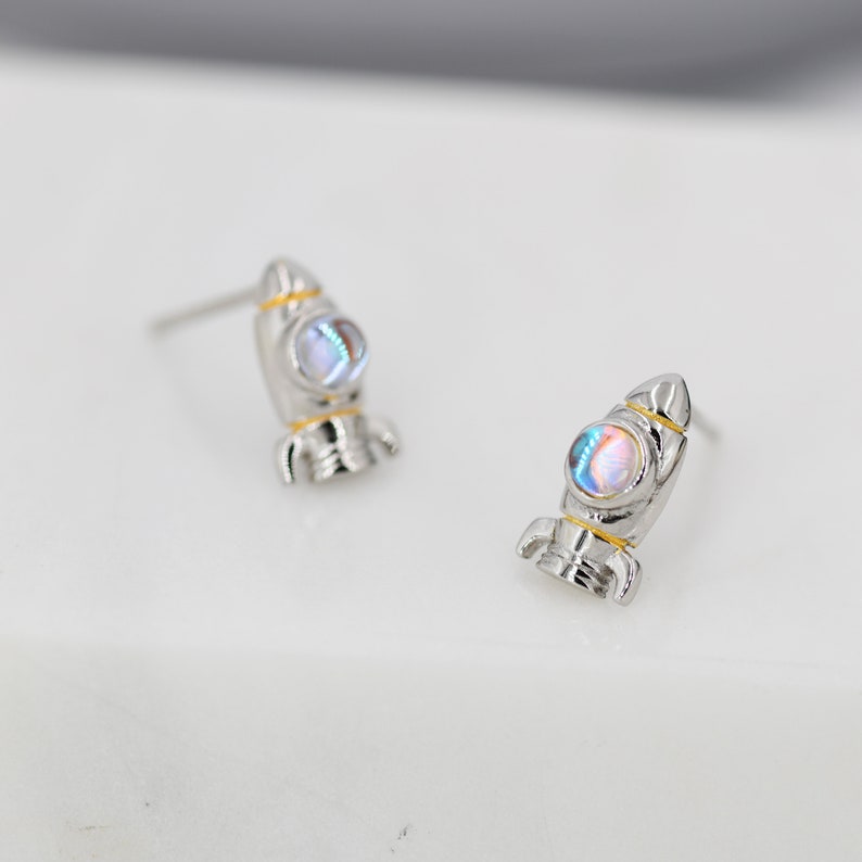 May include: A pair of silver rocket ship earrings with iridescent blue and white stones. The rocket ships have gold accents.