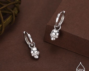 May include: A pair of silver hoop earrings, each featuring a small, polished skull charm dangling from the bottom. The skull charms are detailed with clear, sparkling stones set into the eye sockets, creating a reflective and metallic appearance.