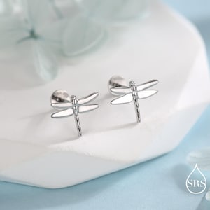 May include: A pair of silver dragonfly stud earrings. Each earring features a detailed dragonfly design with delicate wings and a slender body. The earrings are displayed on a white surface, with a soft, blurred background.