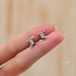 Könnte beinhalten: Ein Paar silberne Ohrringe in Form von Stacheldraht. Die Ohrringe sind klein und zart und perfekt, um jedem Outfit einen Hauch von Coolness zu verleihen.