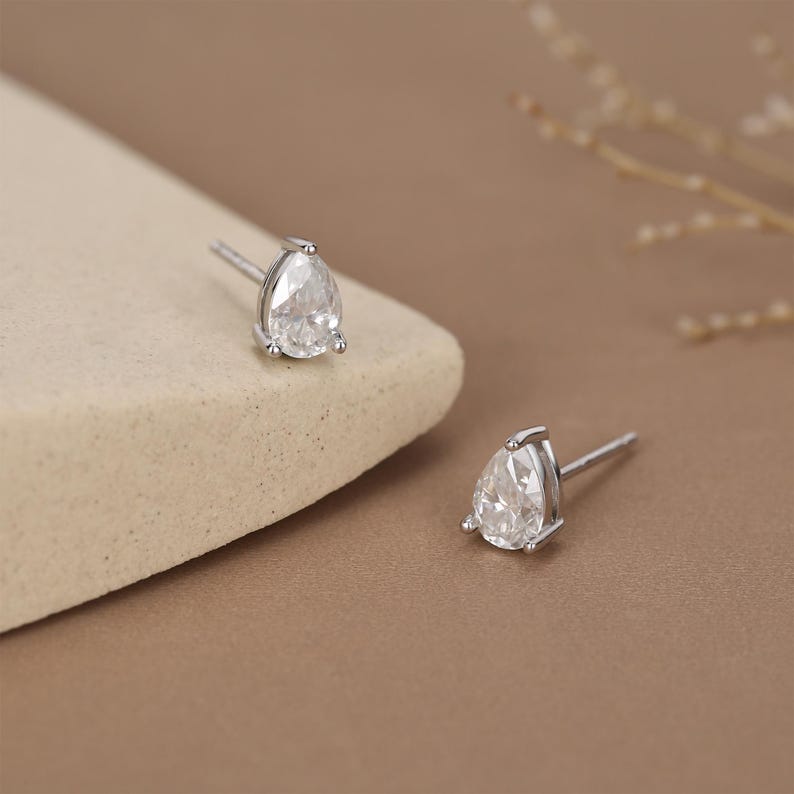May include: A pair of silver stud earrings, each featuring a pear-shaped, clear gemstone. The earrings are set in a simple, elegant design, with a post for pierced ears. The background is a neutral beige and brown.
