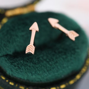 Könnte beinhalten: Ein Paar goldene Pfeilohrringe in einer grünen Samt-Schmuckschatulle. Die Pfeile sind schlicht und zart, mit einer spitzen Spitze und einer kleinen, dreieckigen Befiederung.