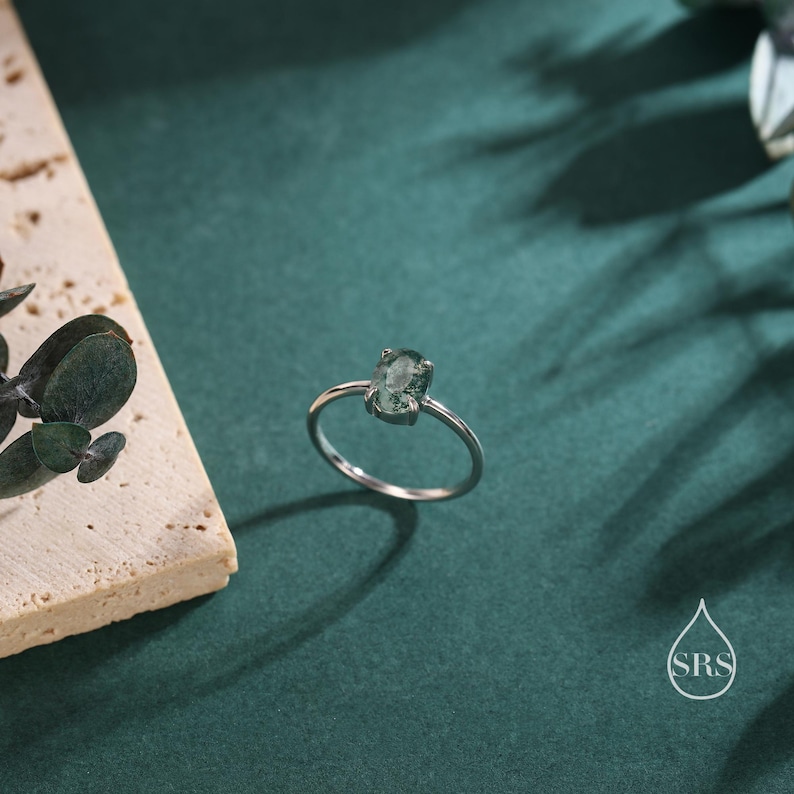 May include: A silver ring with a green gemstone set in a bezel setting. The ring is on a green background.