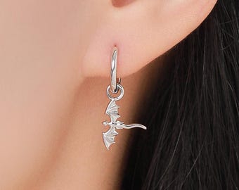 May include: A silver hoop earring featuring a miniature dragon pendant. The dragon is shown with its wings spread, and a long tail. The earring is displayed on a person's ear.