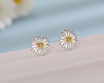 May include: A pair of daisy stud earrings. Each earring features a silver-colored daisy with a yellow center. The petals are detailed and radiate outwards. The earrings are displayed on a light gray surface against a soft blue background.