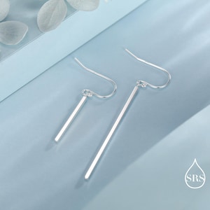 Op de afbeelding: Een paar zilverkleurige oorhangers. Elke oorbel heeft een lange, slanke staaf die aan een gebogen oorhaak hangt. De oorbellen worden gepresenteerd tegen een lichtblauwe achtergrond, met een minimalistisch en modern ontwerp.