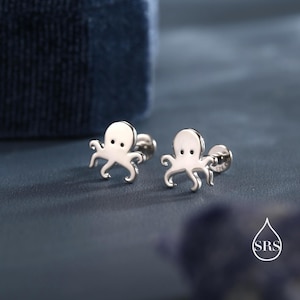 Puede incluir: Un par de pendientes de plata con diseño de pulpo. Cada pendiente presenta un pequeño y detallado diseño de pulpo con una cabeza redondeada y ocho tentáculos. Sobre un fondo azul oscuro.