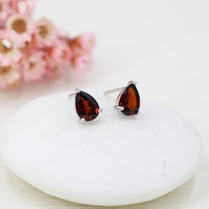 May include: A pair of silver stud earrings, each featuring a teardrop-shaped, deep red garnet gemstone. The earrings are presented on a white surface, with a blurred pink floral arrangement in the background.