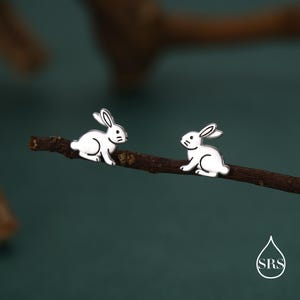 May include: Two white rabbit-shaped earrings with black details are displayed on a brown twig against a dark teal background. The earrings have a minimalist design, with the rabbit's features clearly defined.