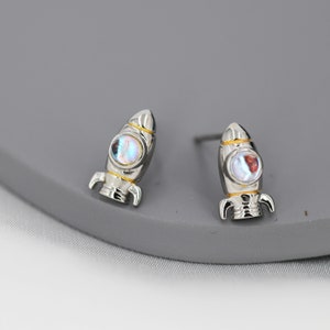 May include: A pair of silver rocket ship stud earrings with iridescent blue and white stones. The rocket ships have gold accents.