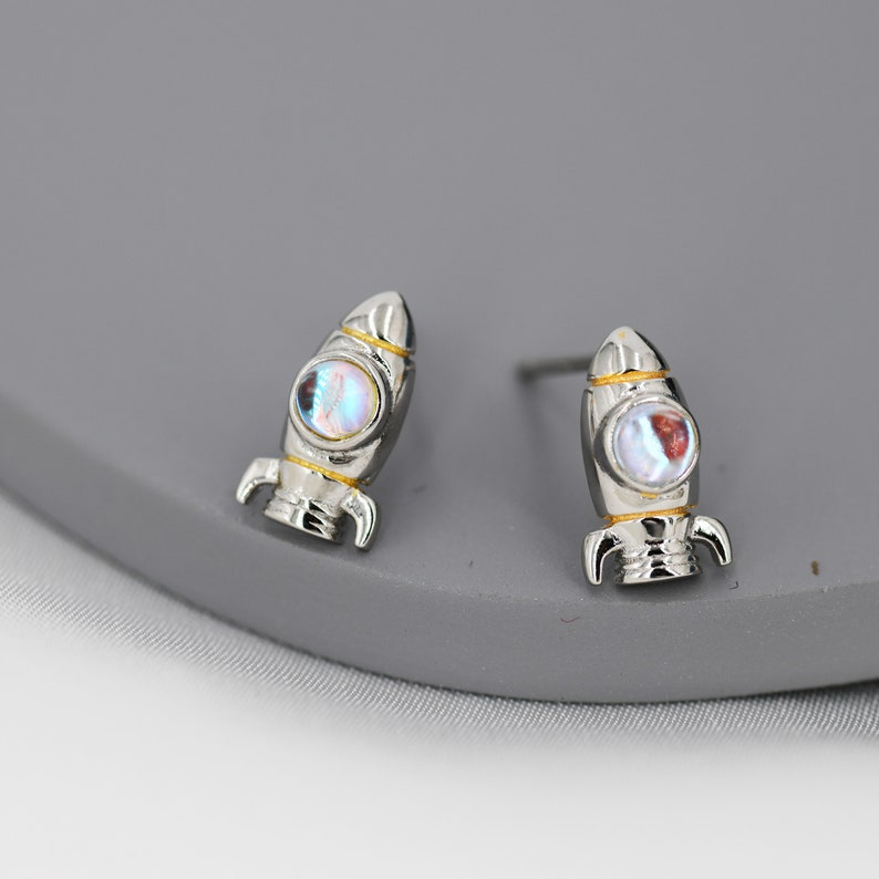 May include: A pair of silver rocket ship stud earrings with iridescent blue and white stones. The rocket ships have gold accents.