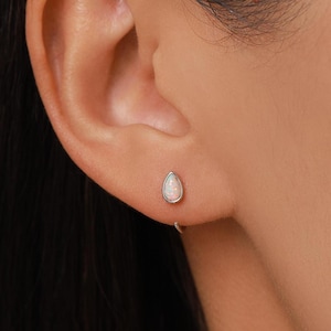 May include: A silver stud earring featuring a teardrop-shaped opal gemstone with iridescent flashes of color. The earring is secured with a metal post and backing, showcasing a smooth, polished silver setting that frames the pear-shaped stone.