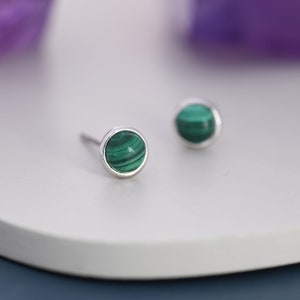 May include: A pair of stud earrings featuring round, polished malachite stones set in a silver bezel. The malachite displays a vibrant green color with distinctive swirling patterns. The earrings are displayed on a white surface.