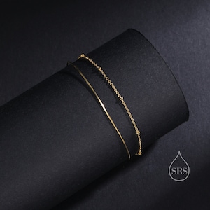 May include: A double-layered gold bracelet featuring one smooth, thin, solid wire band and one delicate chain with small, evenly spaced gold beads. The piece includes a small logo in the corner with a water drop shape and the letters SRS.