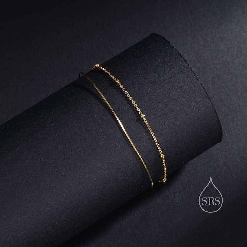 May include: A double-layered gold bracelet featuring one smooth, thin, solid wire band and one delicate chain with small, evenly spaced gold beads. The piece includes a small logo in the corner with a water drop shape and the letters SRS.
