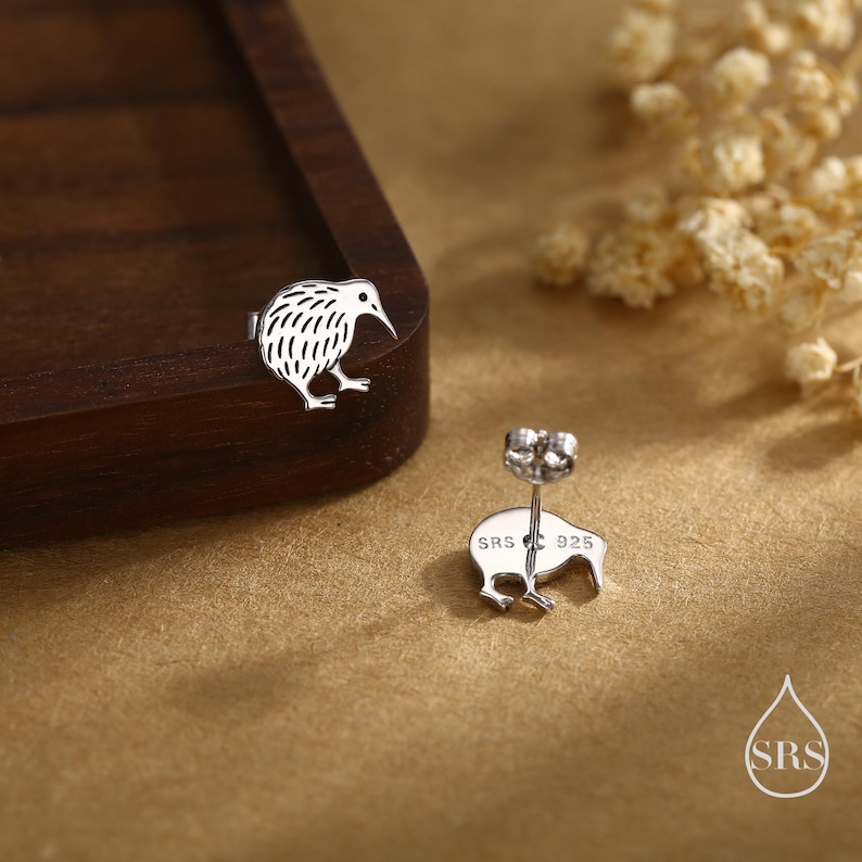 May include: A pair of silver kiwi bird stud earrings. The earrings are detailed with etched feathers and a curved beak. The earring posts are visible, with the stamp "SRS 925". The earrings are displayed on a brown surface with dried flowers.