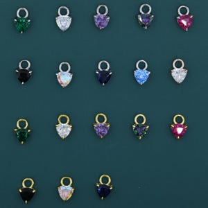 May include: A collection of heart-shaped charms in various colors, including green, clear, purple, iridescent, and pink. Each charm has a small loop at the top for attaching to jewelry. The charms are set against a dark teal background.