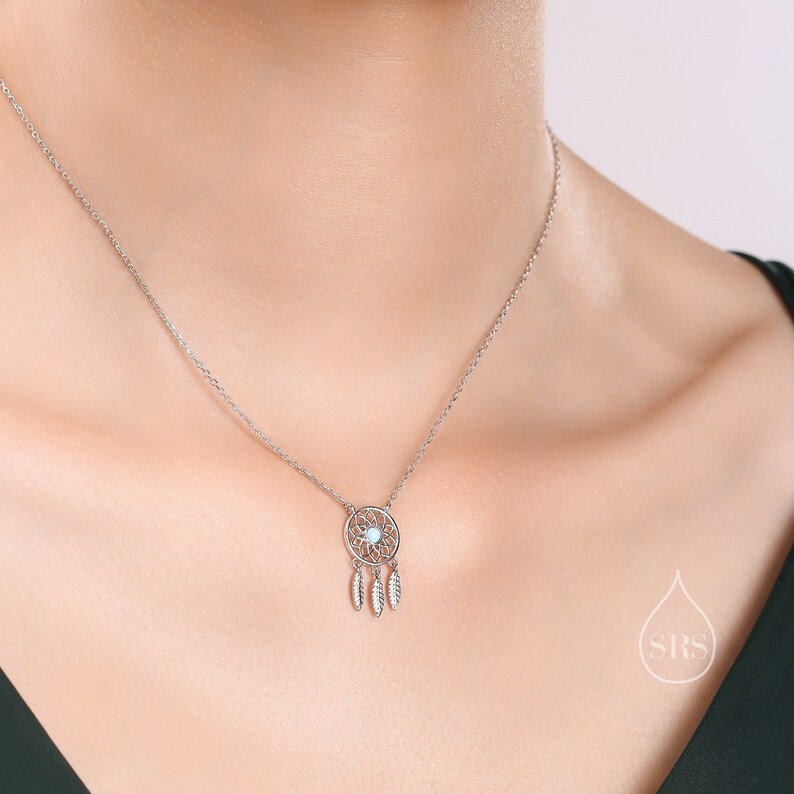 May include: A silver-toned necklace featuring a circular dreamcatcher pendant with a central light blue stone and three small dangling feather charms. The pendant is suspended from a delicate silver-toned chain that rests against the neck.
