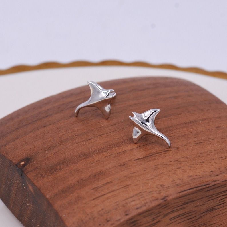 May include: A pair of silver-toned earrings shaped like manta rays. The earrings are small and delicate, and they are perfect for adding a touch of whimsy to any outfit.