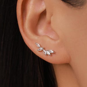 May include: A silver-toned ear climber earring featuring four marquise-cut clear stones arranged in a curved, leaf-like pattern. The earring follows the natural contour of the earlobe, showcasing a bright, reflective metallic finish and faceted gems.