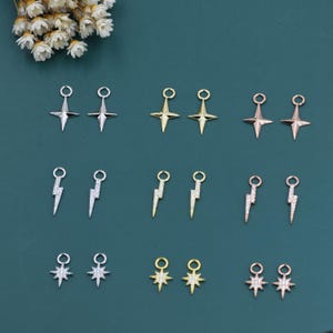 Könnte beinhalten: Verschiedene kleine dekorative Anhänger in Silber, Gold und Roségold. Designs umfassen Sternschnuppen, Blitzbolzen und vierzackige Sterne, jeweils mit einer kleinen Schlaufe. Präsentiert auf einem dunkelblauen Hintergrund.