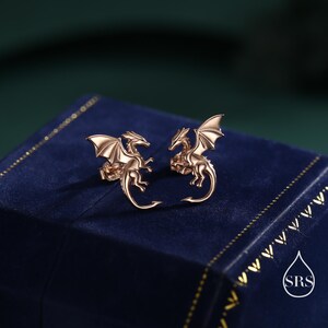 May include: Rose gold dragon stud earrings. Each earring depicts a dragon with detailed wings and a curled tail. The earrings are presented on a dark blue surface with a gold border. A fantasy-themed jewellery item.