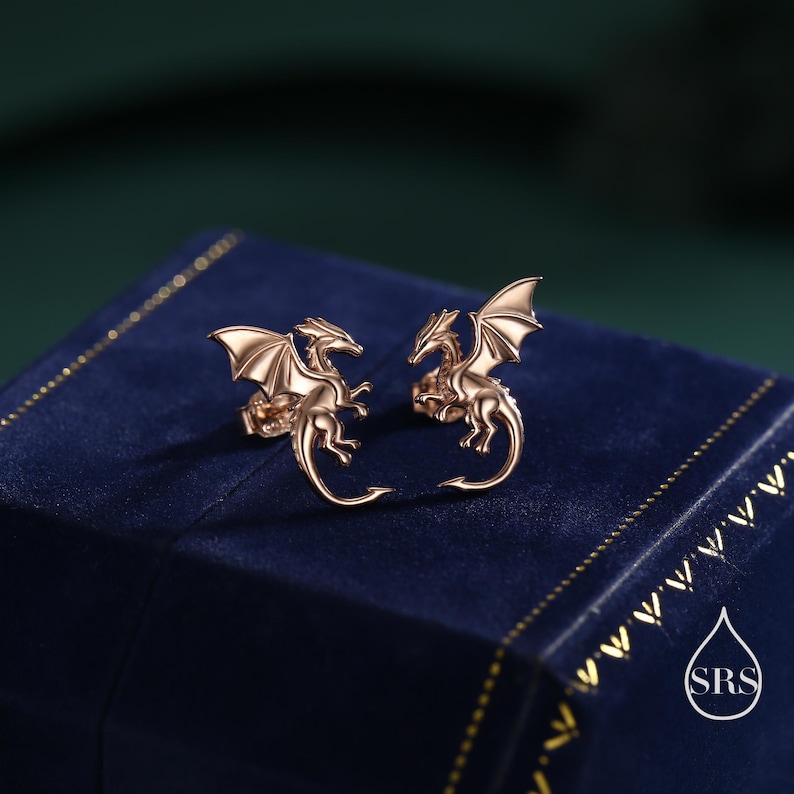 May include: Rose gold dragon stud earrings. Each earring depicts a dragon with detailed wings and a curled tail. The earrings are presented on a dark blue surface with a gold border. A fantasy-themed jewellery item.