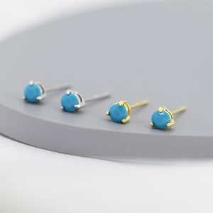 Puede incluir: Un par de pendientes de piedra turquesa con postes de plata y oro. Los pendientes son pequeños y redondos.