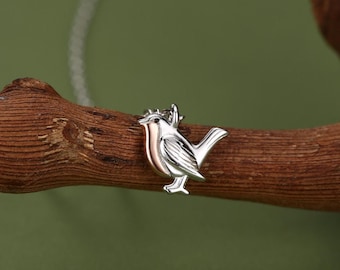 May include: A silver and rose gold bird pendant necklace. The bird is perched on a brown branch against a green background. The bird's chest is rose gold, and the rest of the bird is silver. The necklace chain is also silver.