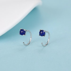 Peut inclure: Une paire de boucles d'oreilles créoles argentées, chacune ornée d'une pierre précieuse ronde bleu foncé. Les boucles d'oreilles ont un design simple et élégant, avec la pierre précieuse placée en haut de la créole. Le fond est bleu clair.