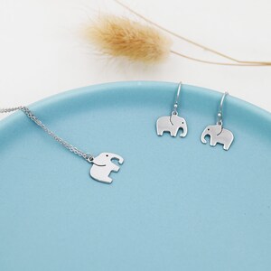 May include: A silver necklace and earrings set featuring a small elephant charm. The jewelry is displayed on a light blue plate.