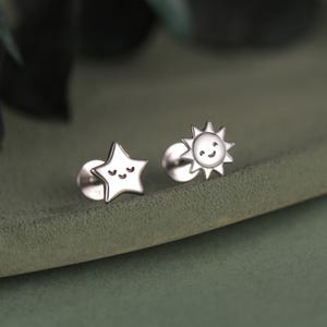 以下が含まれることがあります： 銀色のスタッドピアスのペア。片方のイヤリングは笑顔の星型で、もう片方は笑顔の太陽型です。イヤリングは緑色の表面にあります。