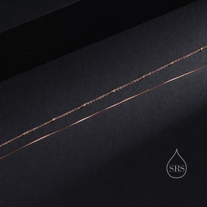 May include: A double-layered rose gold necklace featuring one delicate chain with small evenly spaced beads and a second smooth, thin snake chain. The piece includes a small white logo graphic in the corner with a water drop shape and the letters SRS.