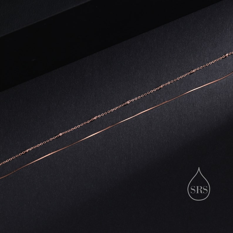 May include: A double-layered rose gold necklace featuring one delicate chain with small evenly spaced beads and a second smooth, thin snake chain. The piece includes a small white logo graphic in the corner with a water drop shape and the letters SRS.