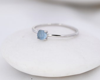May include: A thin silver-toned ring featuring a single, small, round, light blue gemstone set in a four-prong setting. The band is smooth and polished, showcasing a simple design that highlights the pale blue stone as the central focal point.