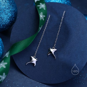 Può includere: Un paio di orecchini pendenti in argento con un ciondolo a forma di manta. Gli orecchini sono appesi su uno sfondo blu con un nastro verde decorato con fiocchi di neve bianchi.
