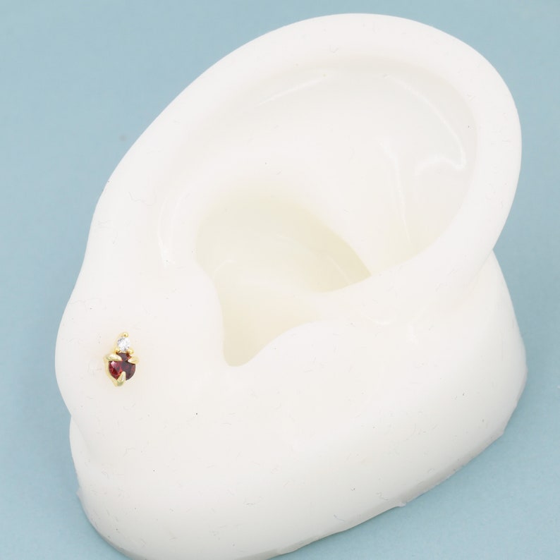 May include: A close-up of a red gemstone nose ring with a small clear gemstone accent. The ring is on a white ear model.