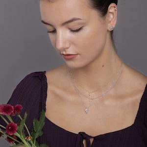 May include: A layered necklace set featuring a gold-tone chain with a lightning bolt pendant and a silver-tone chain with a small sword charm. The jewelry is worn against a dark purple top, complemented by a small silver stud earring in the earlobe.