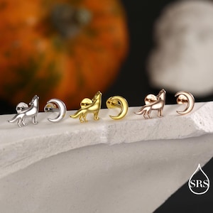 Op de afbeelding: Stud oorbellen met een huilende wolf en maan ontwerp. Verkrijgbaar in zilver, goud en roségoud. Gepresenteerd op een wit oppervlak met een vage achtergrond. Sieraden voor doorboorde oren.