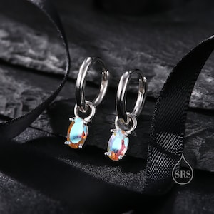 Può includere: Un paio di orecchini a cerchio d'argento con una pietra preziosa ovale iridescente pendente. Gli orecchini sono su una superficie nera.