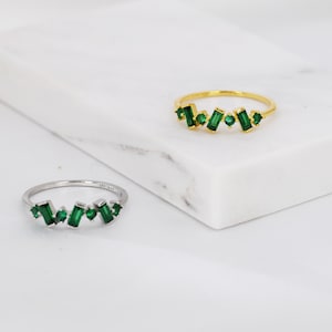 Könnte beinhalten: Zwei Ringe mit einer Reihe grüner Smaragdschliffsteine, die in Gold und Silber gefasst sind.